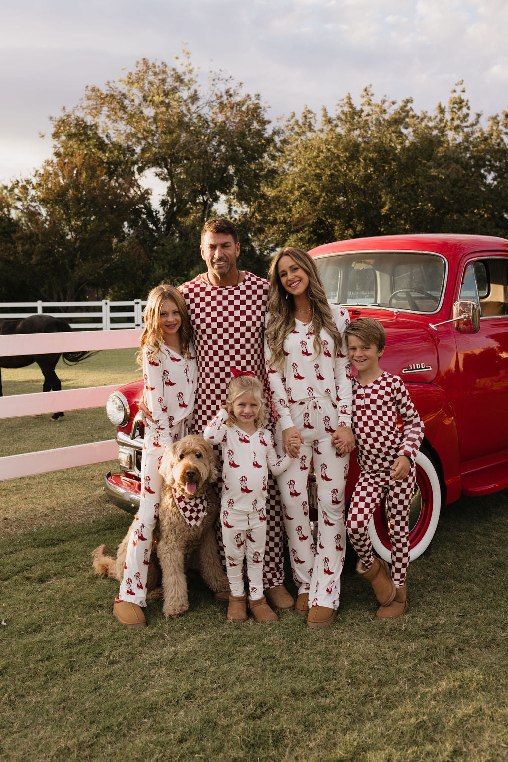 A family of five, dressed in matching Bamboo Two Piece Pajamas from lolo webb's "Cowgirl Christmas" collection, poses outdoors with a playful dog and a vintage red truck. The scene is staged on a grassy area lined with a white fence and surrounded by trees, encapsulating a cozy yet stylish atmosphere.