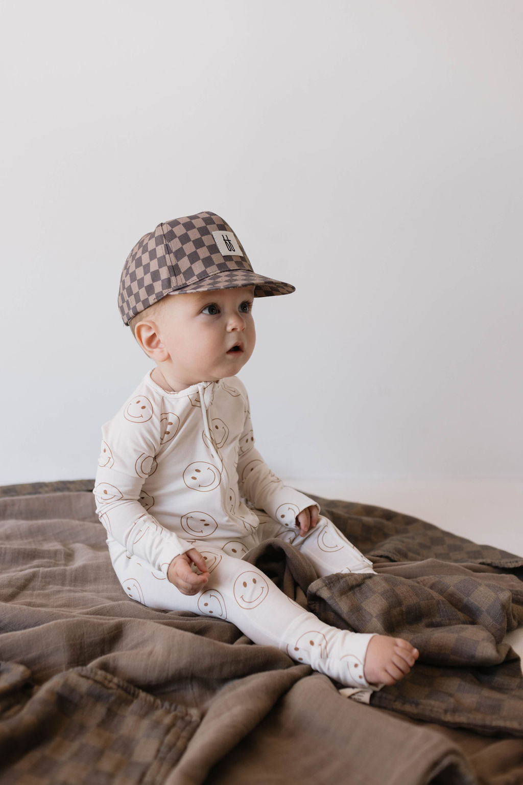 A baby, dressed in a patterned onesie adorned with smiley faces and sporting a forever french baby's Children's Trucker Hat in Faded Brown Checkerboard that provides sun protection, sits on a cozy quilt. The baby looks curiously to the side against a neutral, light-colored backdrop.