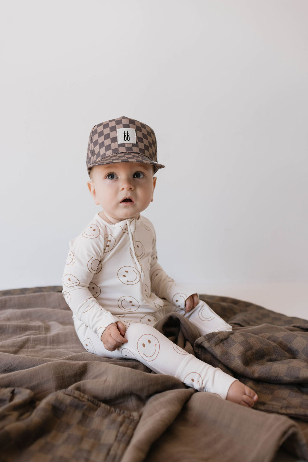 A baby in a white outfit adorned with smiley faces sits on a brown blanket. The baby is wearing the "Children's Trucker Hat | Faded Brown Checkerboard" by forever french baby, perfect for sun protection, and looks towards the camera with an expression of curiosity. The background is plain white.