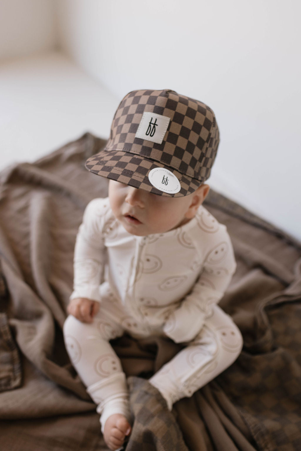 A baby wearing the forever french baby's Children's Trucker Hat in Faded Brown Checkerboard, providing stylish sun protection, and a white onesie with a paisley pattern sits on a brown blanket. The baby appears curious, looking up towards the camera in this minimalistic setting with a neutral-colored background.