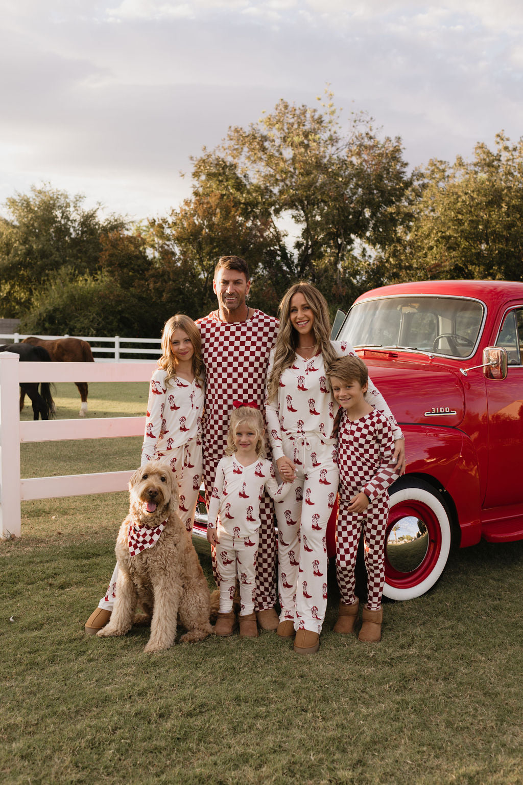 A family of five and a dog, all dressed in matching Bamboo Two Piece Pajamas from lolo webb's Cowgirl Christmas collection, stand outside by a vintage red truck. They are smiling against the backdrop of a horse and trees.