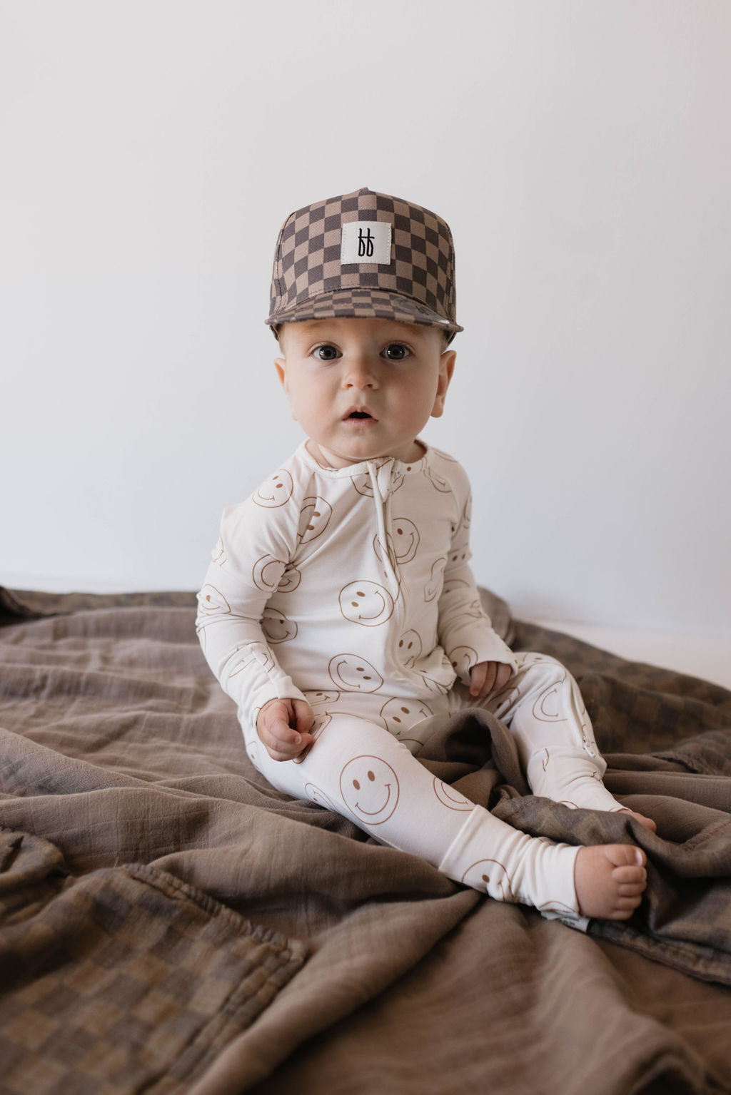 A baby in a one-piece outfit adorned with smiley faces sits on a brown blanket. To keep the sun at bay, the baby is sporting an adorable Children's Trucker Hat in Faded Brown Checkerboard by forever french baby. The background is a plain, light-colored wall.