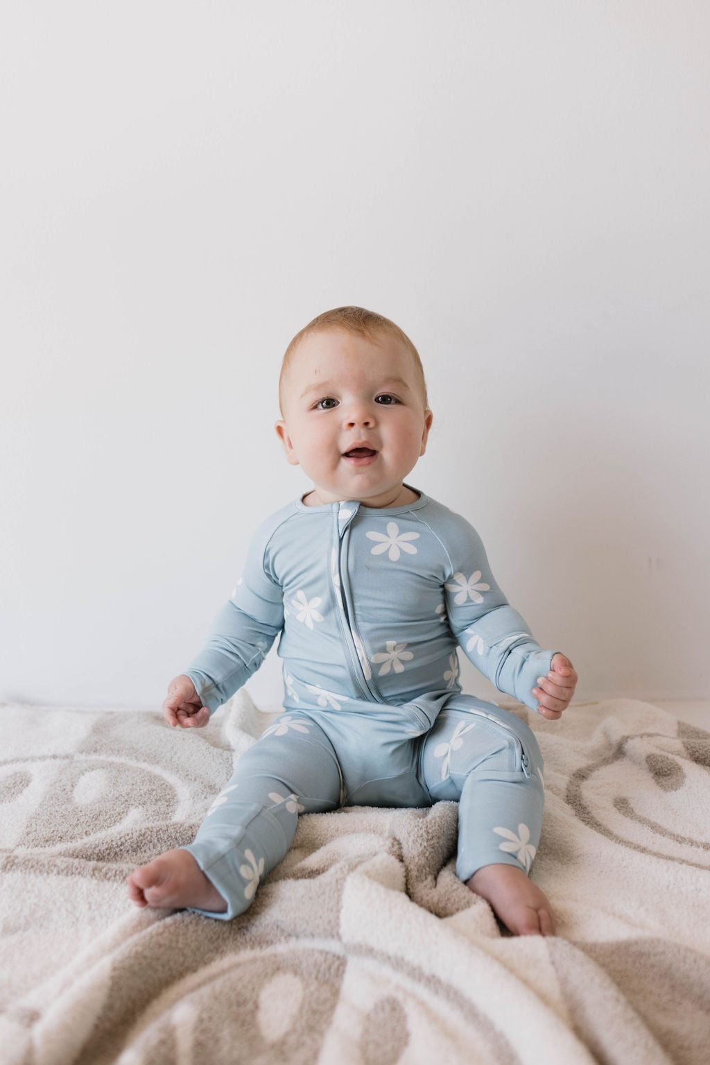 A baby, dressed in forever french baby's Bamboo Zip Pajamas in the Daisy Blues pattern, sits on a soft, patterned blanket. The baby has an open-mouthed expression and is looking slightly upward against a plain white wall backdrop.