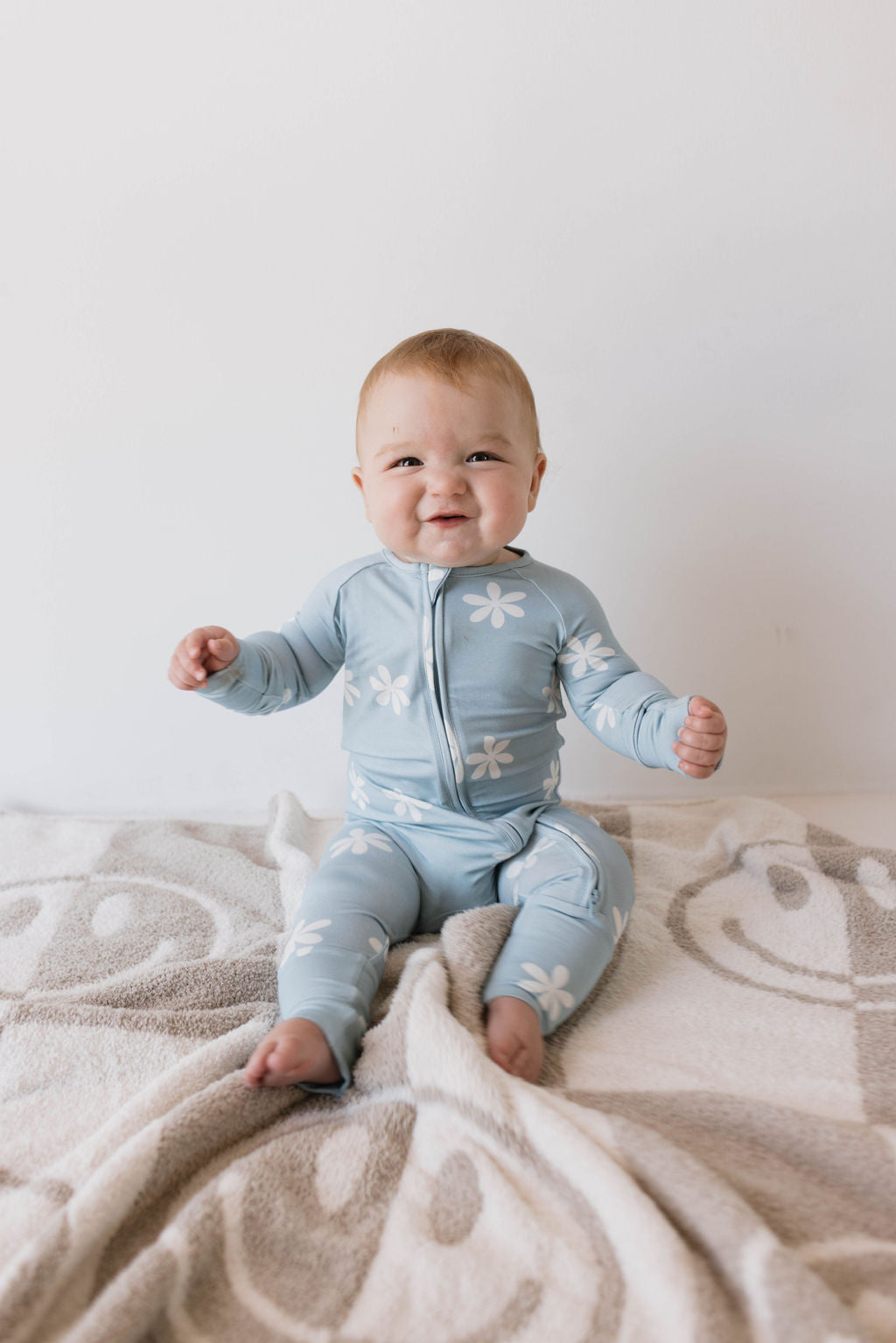 A baby wearing Bamboo Zip Pajamas in the Daisy Blues pattern by forever french baby is sitting on a blanket that features smiling faces. The baby is smiling, has light hair, both arms slightly raised, and is barefoot.