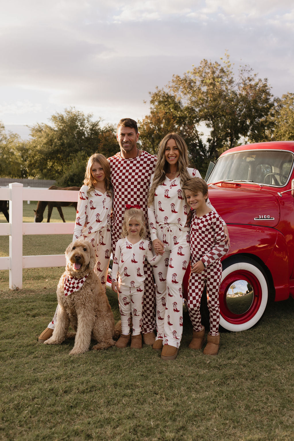 Dressed in their Cowgirl Christmas bamboo pajamas by lolo webb, a family of five poses in front of a red vintage truck. Standing on the grass near a white fence with two horses behind them, their large dog also wears the breathable sleepwear as it sits beside them.