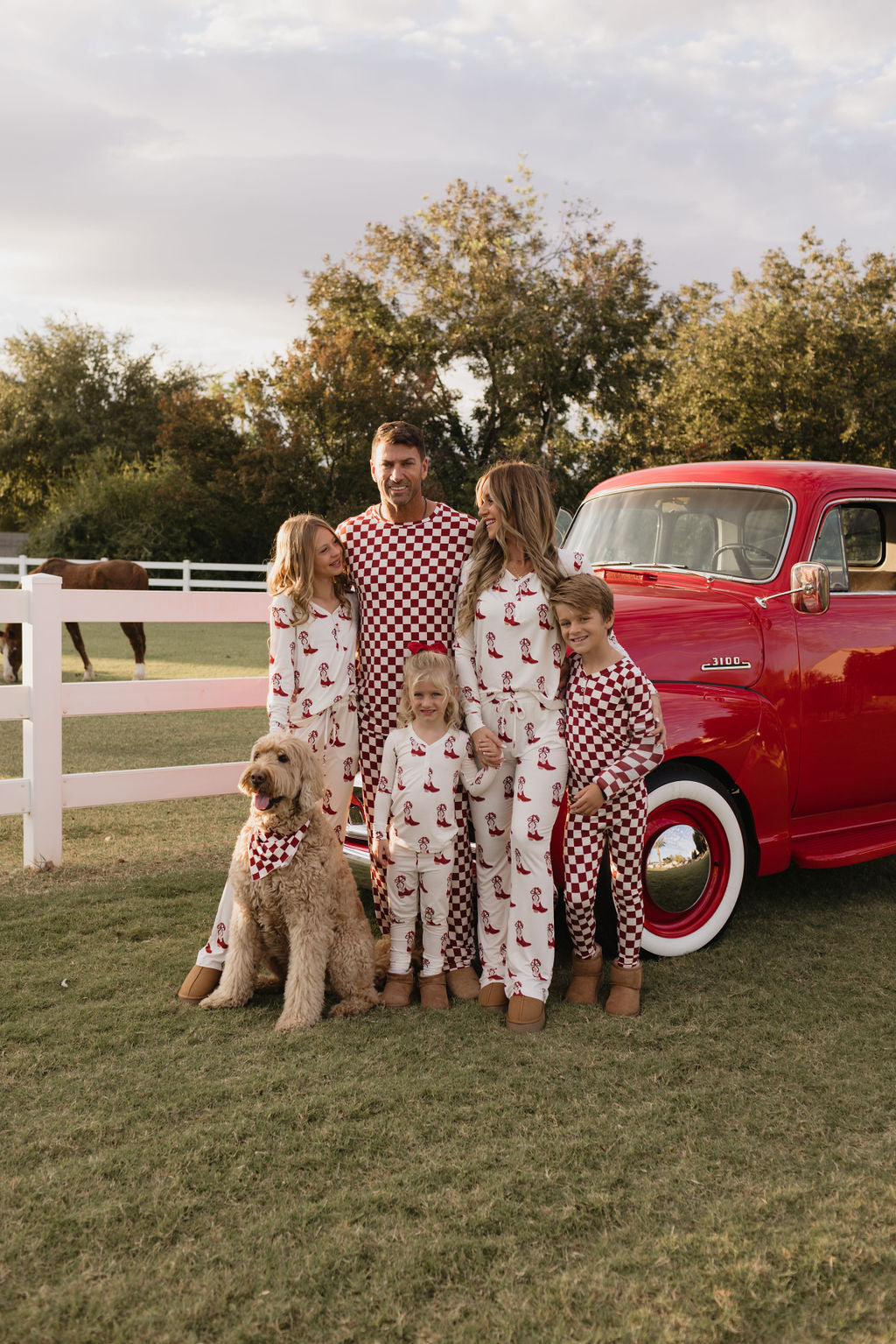 A family stands outdoors, dressed in matching lolo webb Bamboo Two Piece Pajamas in the Cowgirl Christmas pattern. They are beside a red vintage truck, with a dog wearing a matching bandana sitting next to them. A horse grazes in a fenced field nearby under a partly cloudy sky.