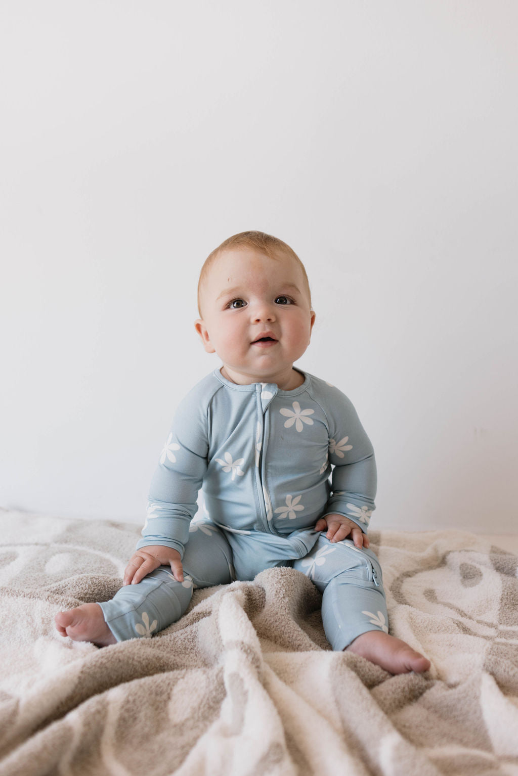 A baby in light blue Bamboo Zip Pajamas from forever french baby, featuring the Daisy Blues pattern, sits on a soft beige blanket. The curious-looking baby gazes slightly upward against a plain white background.