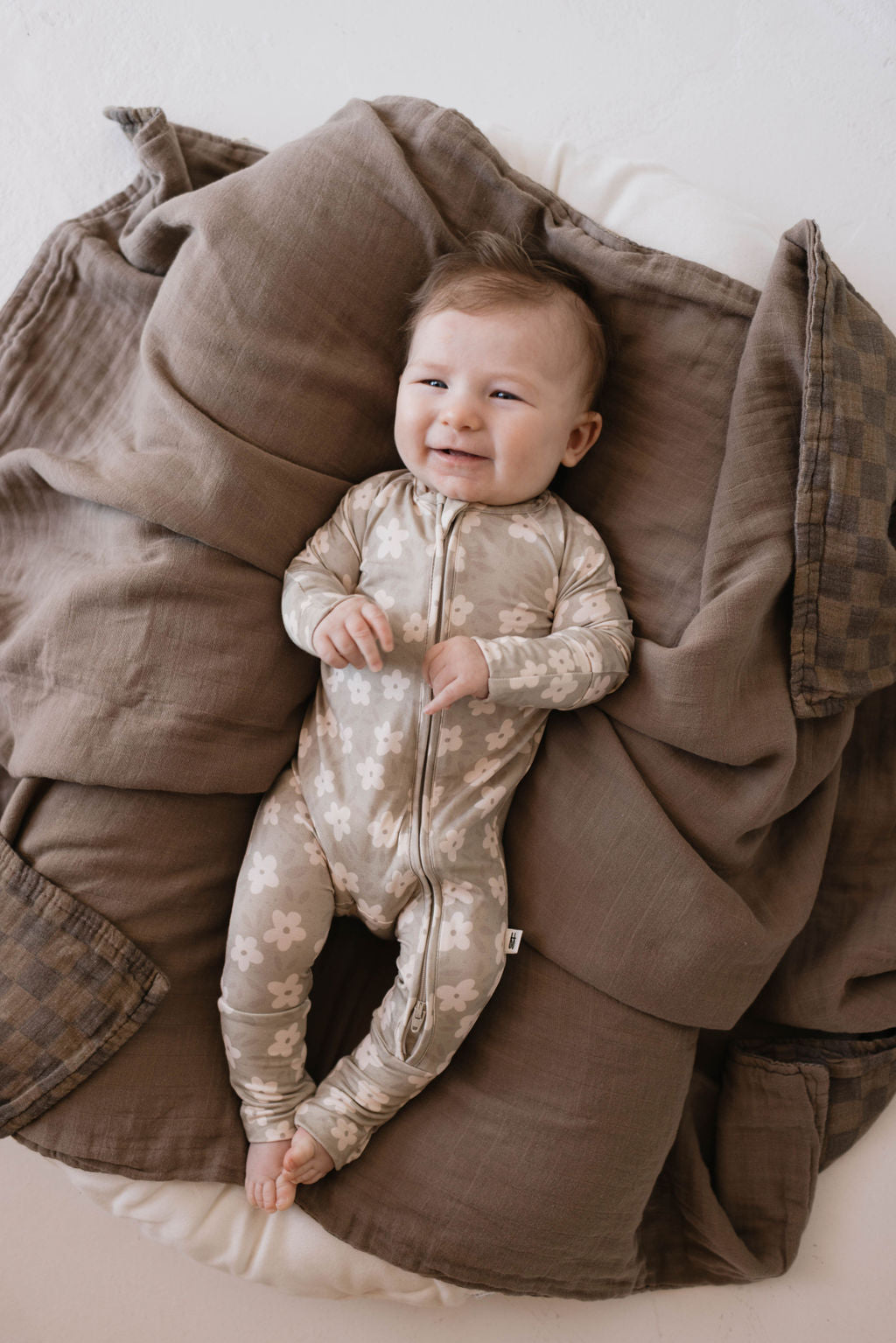 A baby wearing Forever French Baby's Bamboo Zip Pajamas in French Gray Floral smiles while lying on a pile of brown blankets. The baby has light-colored hair and is positioned centrally in the image. The soft blankets provide a warm and comfortable setting, highlighting the comfort of these premium bamboo pajamas.