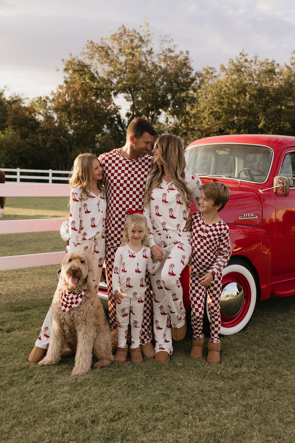 A family of five, plus their dog, is wearing matching lolo webb's Cowgirl Christmas bamboo pajamas. They are gathered beside a vintage red truck in a grassy area surrounded by trees, creating a cozy and joyful scene.