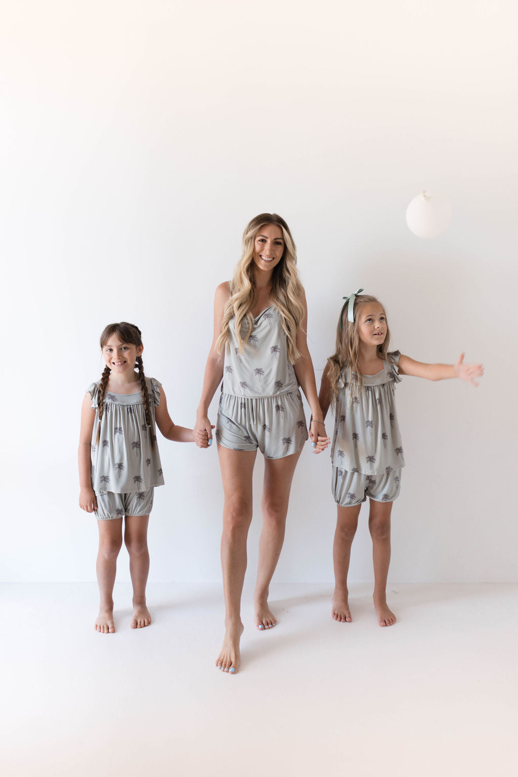 A woman and two young girls stand barefoot in matching Cami Women's Bamboo Sets | Summer Dreamin' from Forever French, made from hypo-allergenic bamboo fabric with small prints. The woman holds hands with both girls, who are smiling. One girl has her arms by her sides, while the other has one arm extended outward. They stand against a plain white background.