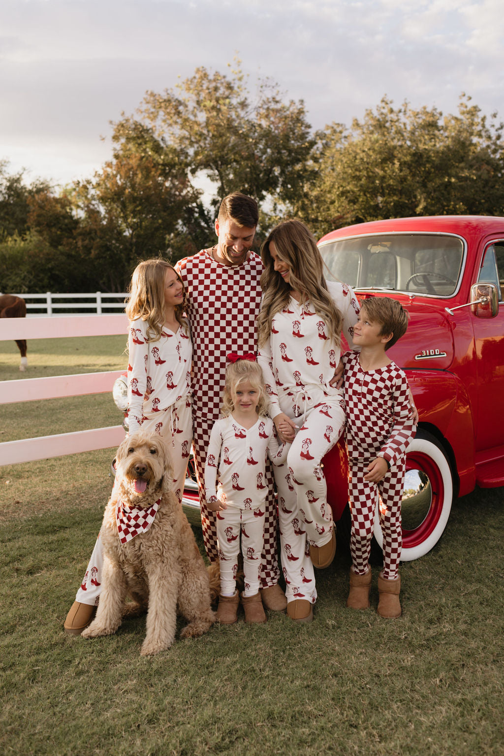 A family of five clad in matching Bamboo Two Piece Pajamas from the Cowgirl Christmas collection by lolo webb stands outdoors next to a vintage red truck, while their large dog joins in with a matching scarf. The scene is beautifully framed by trees and a white fence, providing a picturesque backdrop for their joyful gathering.