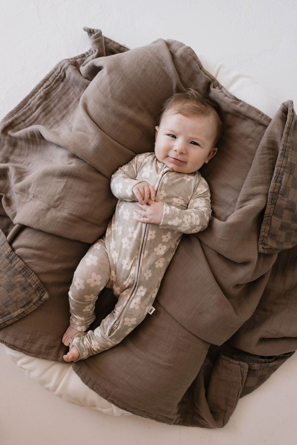 A baby lies on a soft, textured blanket wearing cozy Bamboo Zip Pajamas in French Gray Floral by forever french baby. The baby is looking up and smiling slightly, with their hands gently clasped near their chest. The blanket surrounding them has a neutral, earthy tone.