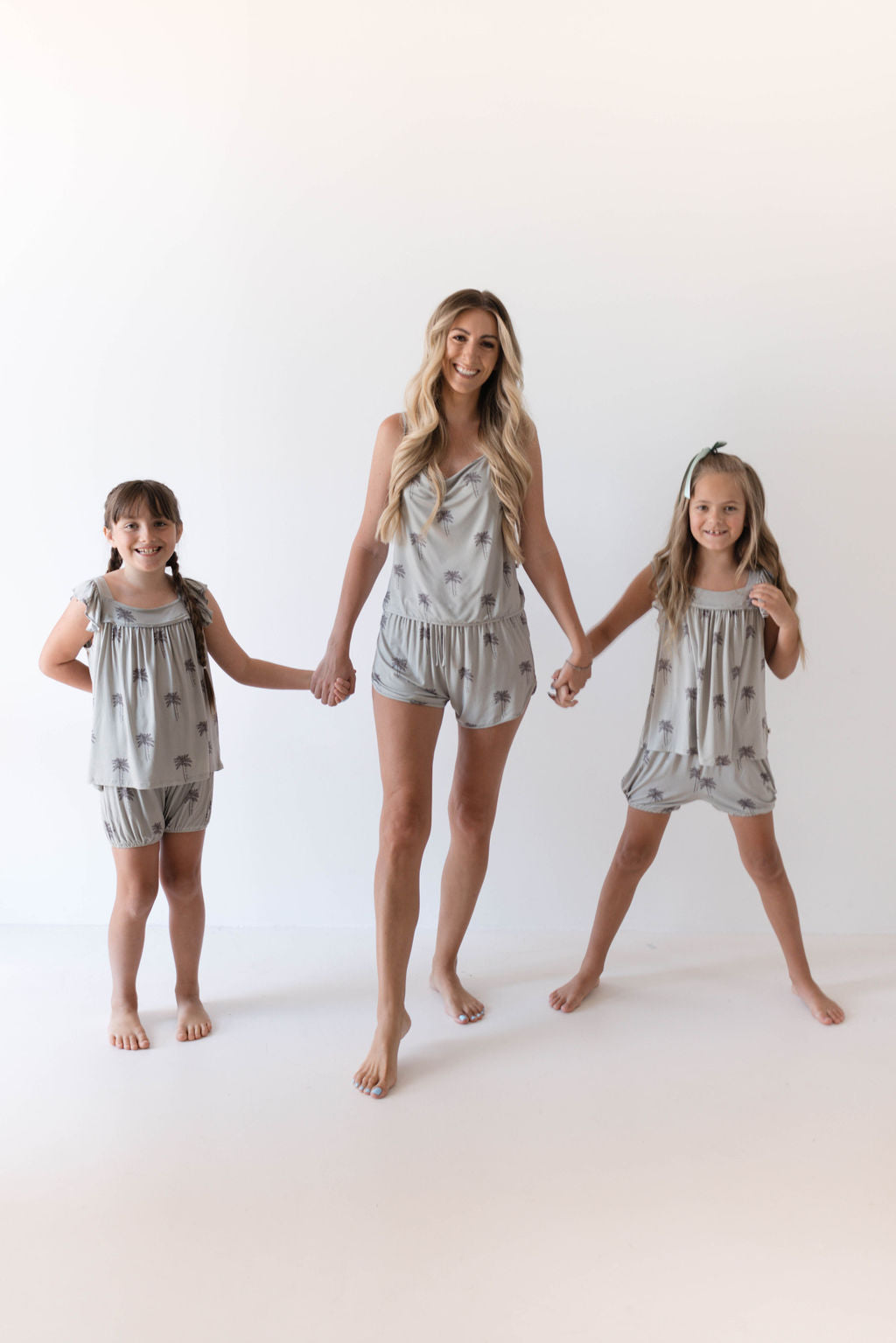 A woman and two young girls stand on a light background, holding hands and smiling. They all wear matching light gray sleeveless outfits made of bamboo fabric with a subtle pattern. The woman is in the middle, and the girls are on either side of her. They appear happy and relaxed in their Forever French Summer Dreamin' Cami Women's Bamboo Set.