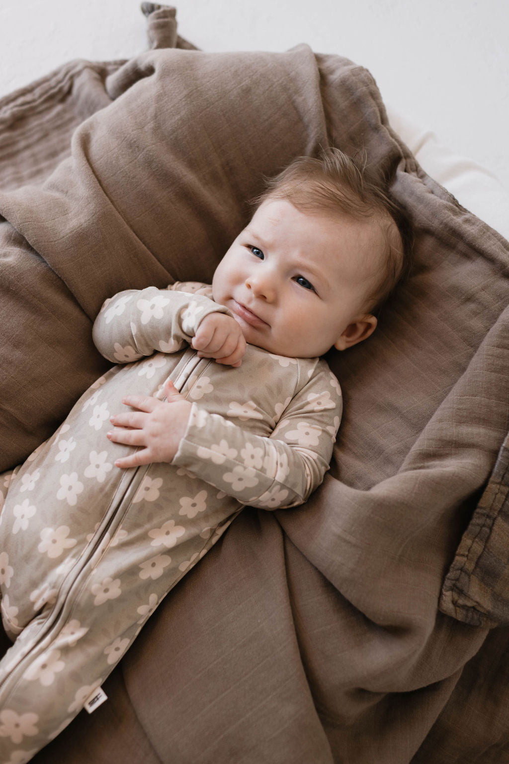 A baby lies on a cozy, brown blanket, dressed in forever french baby's Bamboo Zip Pajamas in French Gray Floral. The baby has a slight smile and appears relaxed, resting one hand on their chest and the other near their mouth. The background is soft and neutral-toned.