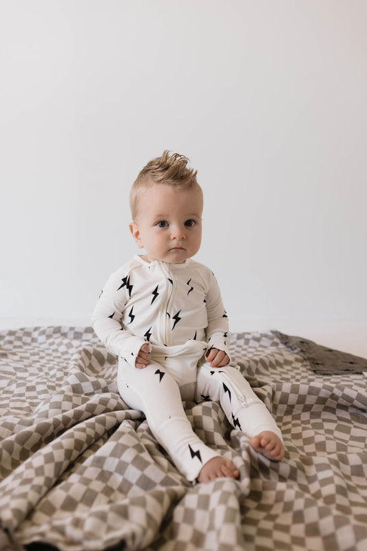 A baby with blond hair, wearing Forever French Baby Bamboo Zip Pajamas in a white and black lightning bolt pattern, sits on a gray and white checkered blanket against a plain background. The baby looks at the camera with a neutral expression.