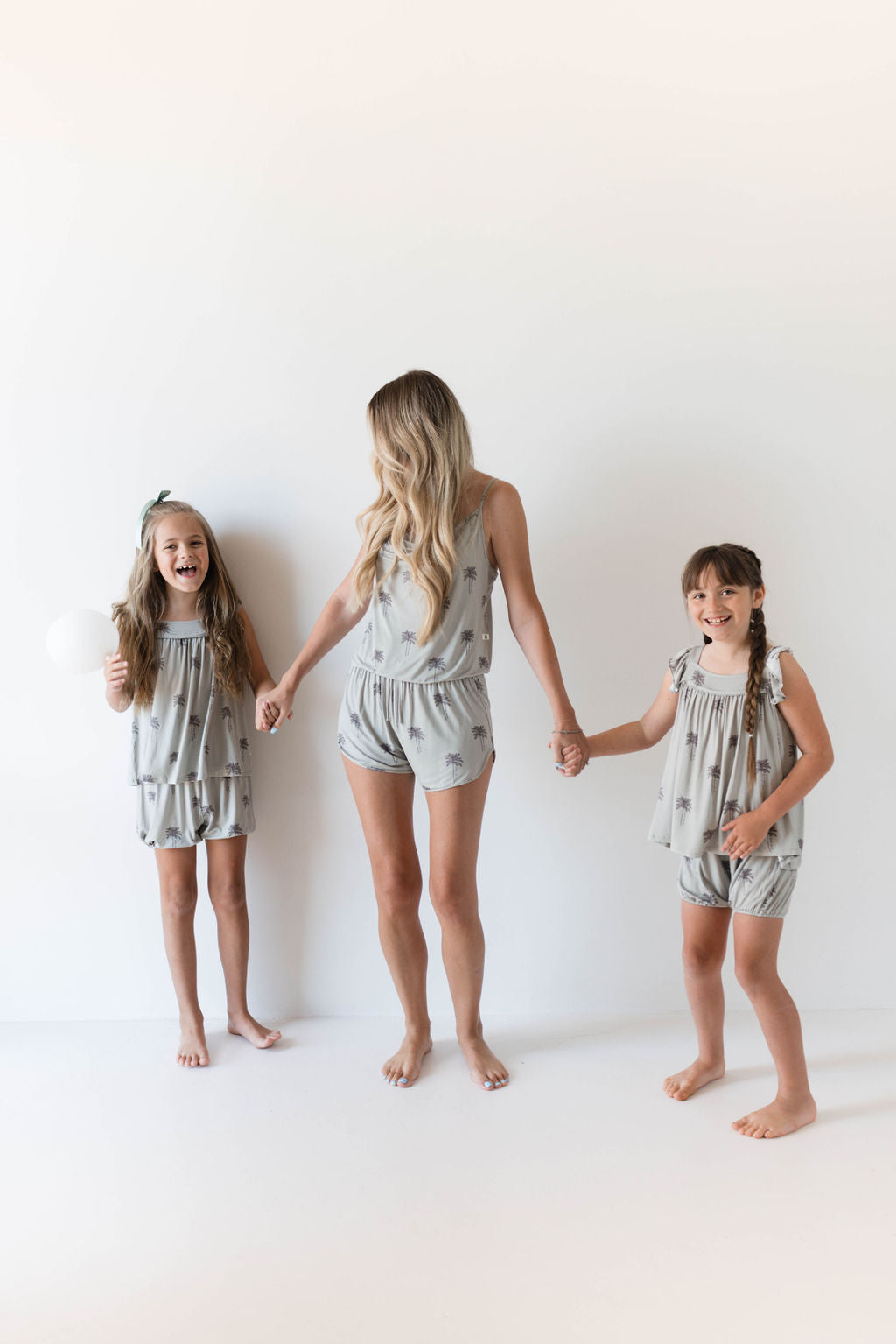 A woman with long blonde hair is holding hands with two young girls. All three are barefoot and wearing matching grey outfits made from hypo-allergenic bamboo fabric. The girl on the left is holding a white balloon. They are standing against a plain white background, showcasing the Cami Women's Bamboo Set | Summer Dreamin' by Forever French.