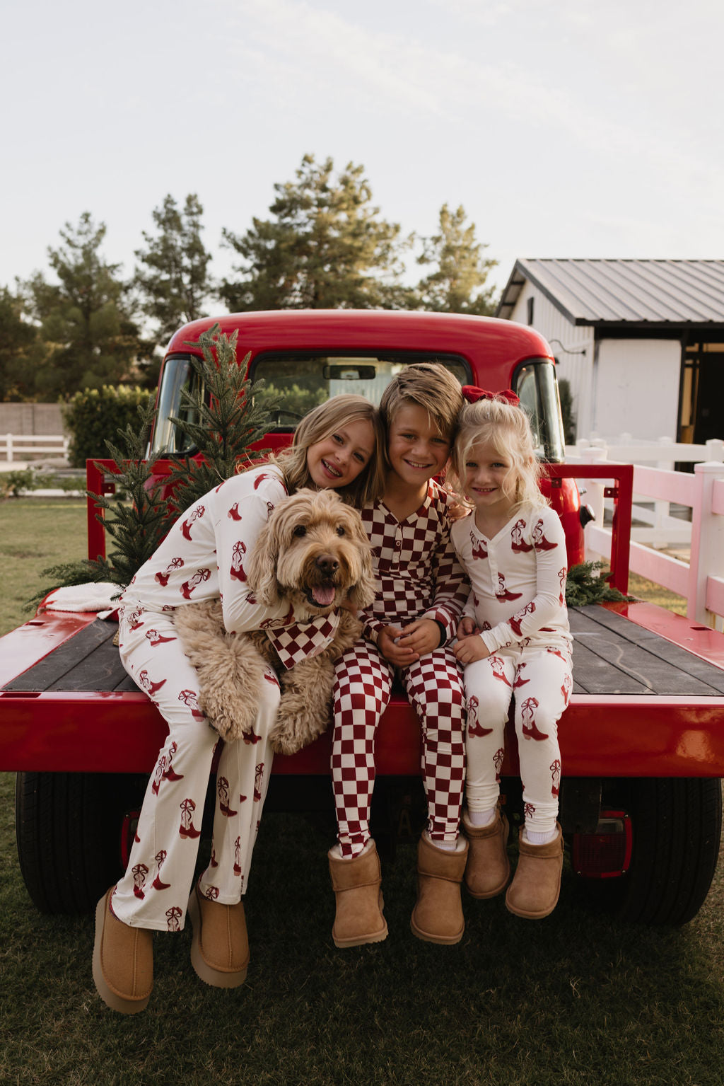 Three children, dressed in matching lolo webb Pre-Teen Flare Bamboo Pajamas in the Cowgirl Christmas design, sit on the back of a red truck with a fluffy dog. They are smiling and wearing cozy boots. A small Christmas tree is in the truck, with a white fence and trees in the background.