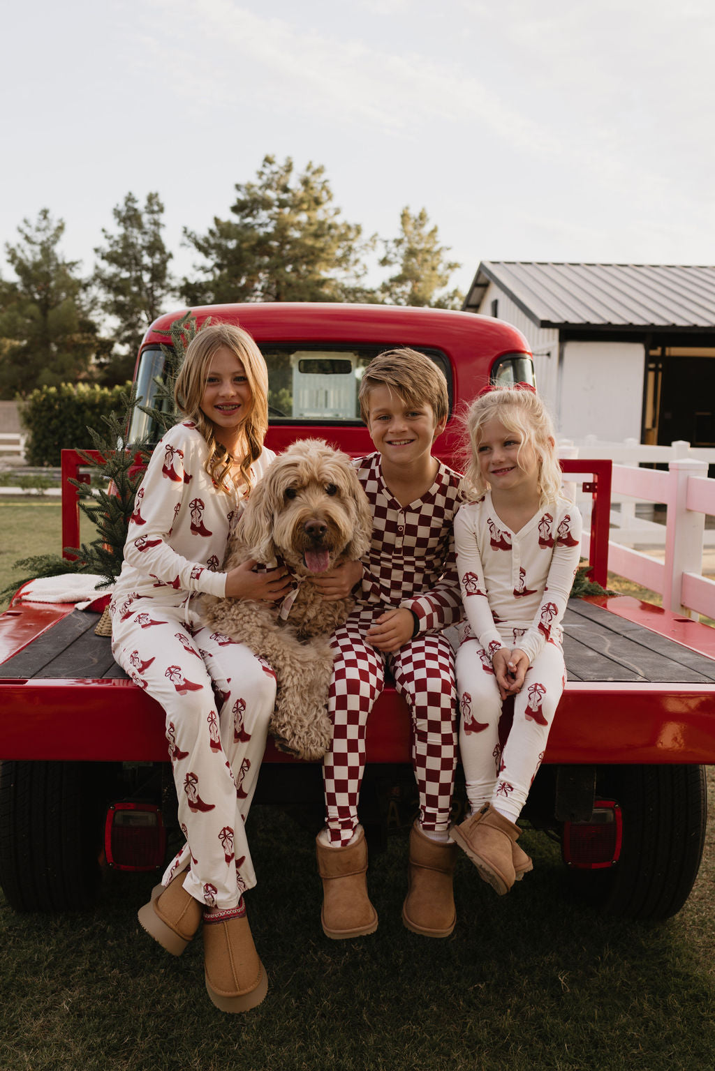 Three children wearing the lolo webb Bamboo Two Piece Pajamas in the Cowgirl Christmas design sit on the back of a red truck, smiling next to a fluffy dog. They are outdoors with trees and a white building in the background, enjoying the cozy comfort of their breathable bamboo apparel.