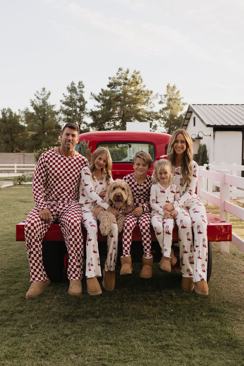 A family of five relaxes on the tailgate of a red truck, all dressed in matching Pre-Teen Flare Bamboo Pajamas from lolo webb’s Cowgirl Christmas collection. The pajamas feature red checker patterns and festive designs made from breathable fabric. A light brown dog sits alongside them, with a barn and trees visible in the background.