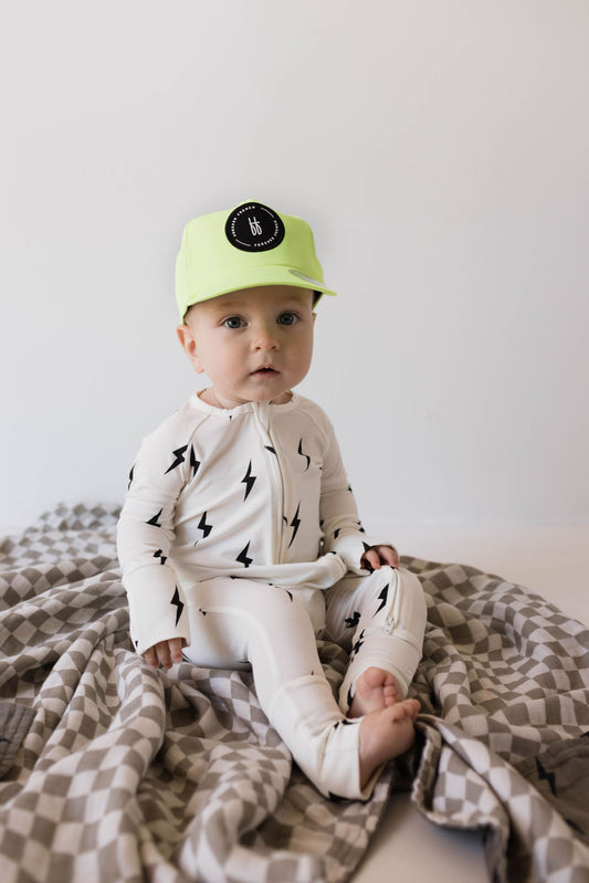 A baby wearing Forever French Baby's Bamboo Zip Pajamas in a white and black lightning bolt design, along with a lime green hat featuring a circular black logo, sits on a grey and white checkered blanket against a plain light background. The child looks directly at the camera with a neutral expression.