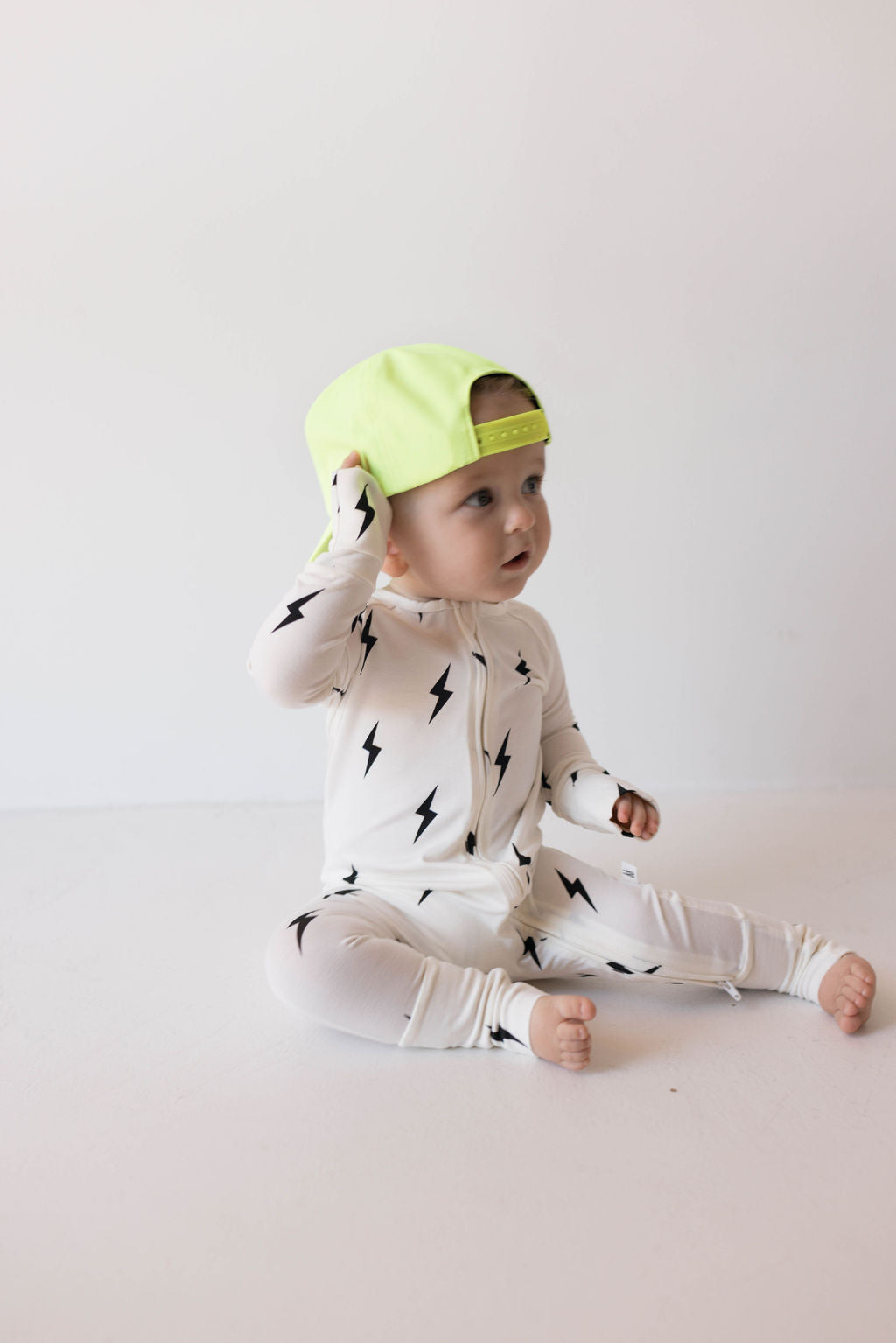 A baby wearing a white onesie with black lightning bolt patterns and the "Children's Trucker Hat | Highlights" by forever french baby sits on the floor. The adjustable fit cap is worn backward as the baby looks to the side, one hand touching it. The plain white background hints at future outdoor adventures.