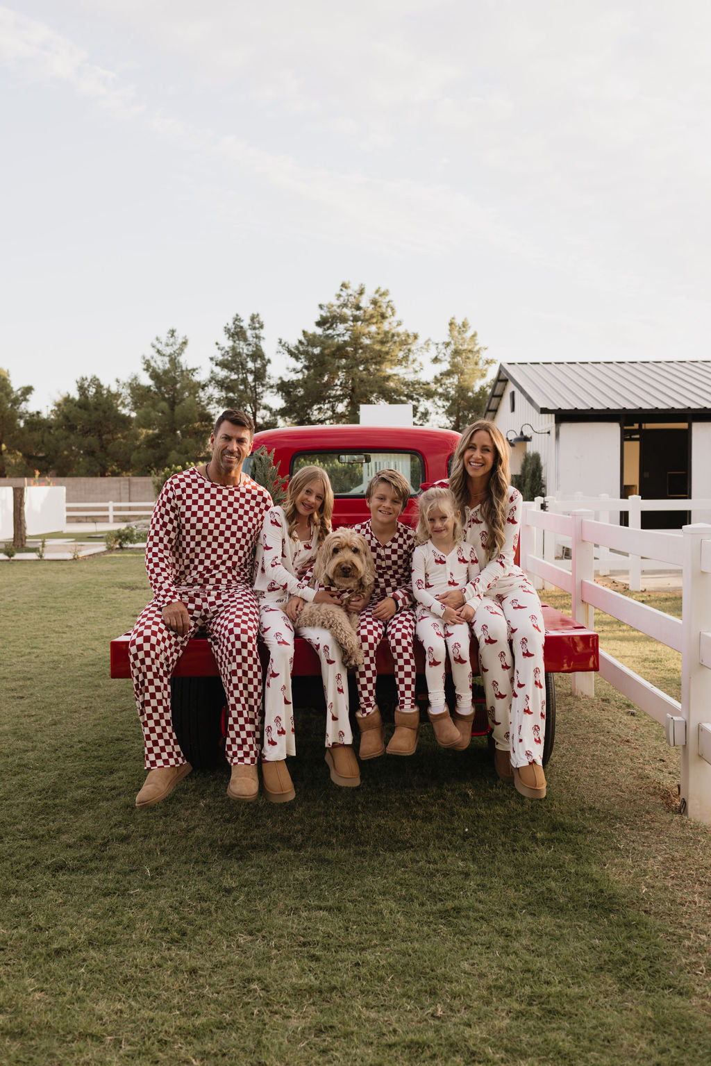 A family of five, dressed in matching lolo webb Pre-Teen Flare Bamboo Pajamas in the Cowgirl Christmas design, relaxes on the tailgate of a red truck. They are outdoors, surrounded by trees and a white building in the background. A fluffy dog joins them as they enjoy each other's company under the clear, bright sky that enhances their cozy gathering.