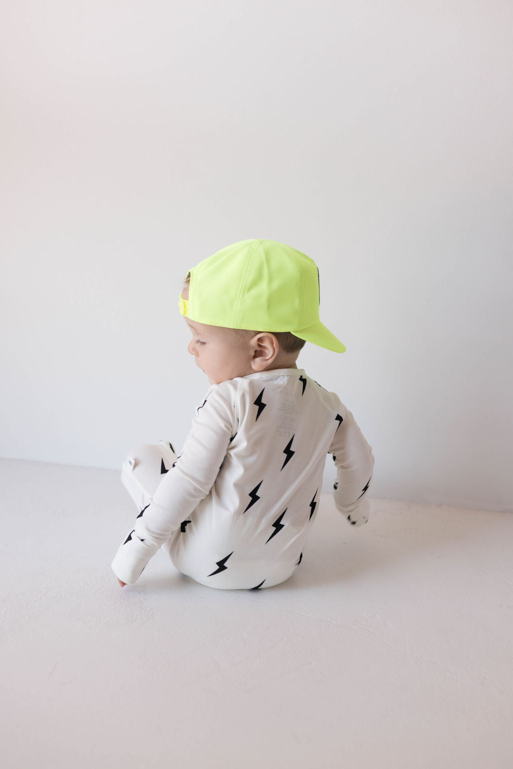 A baby sits on a floor, facing away from the camera, wearing a white onesie with black lightning bolt designs and a neon yellow "Children's Trucker Hat | Highlights" by forever french baby turned backward. The adjustable fit of the cap ensures comfort for all their outdoor adventures. The background is a plain white wall.