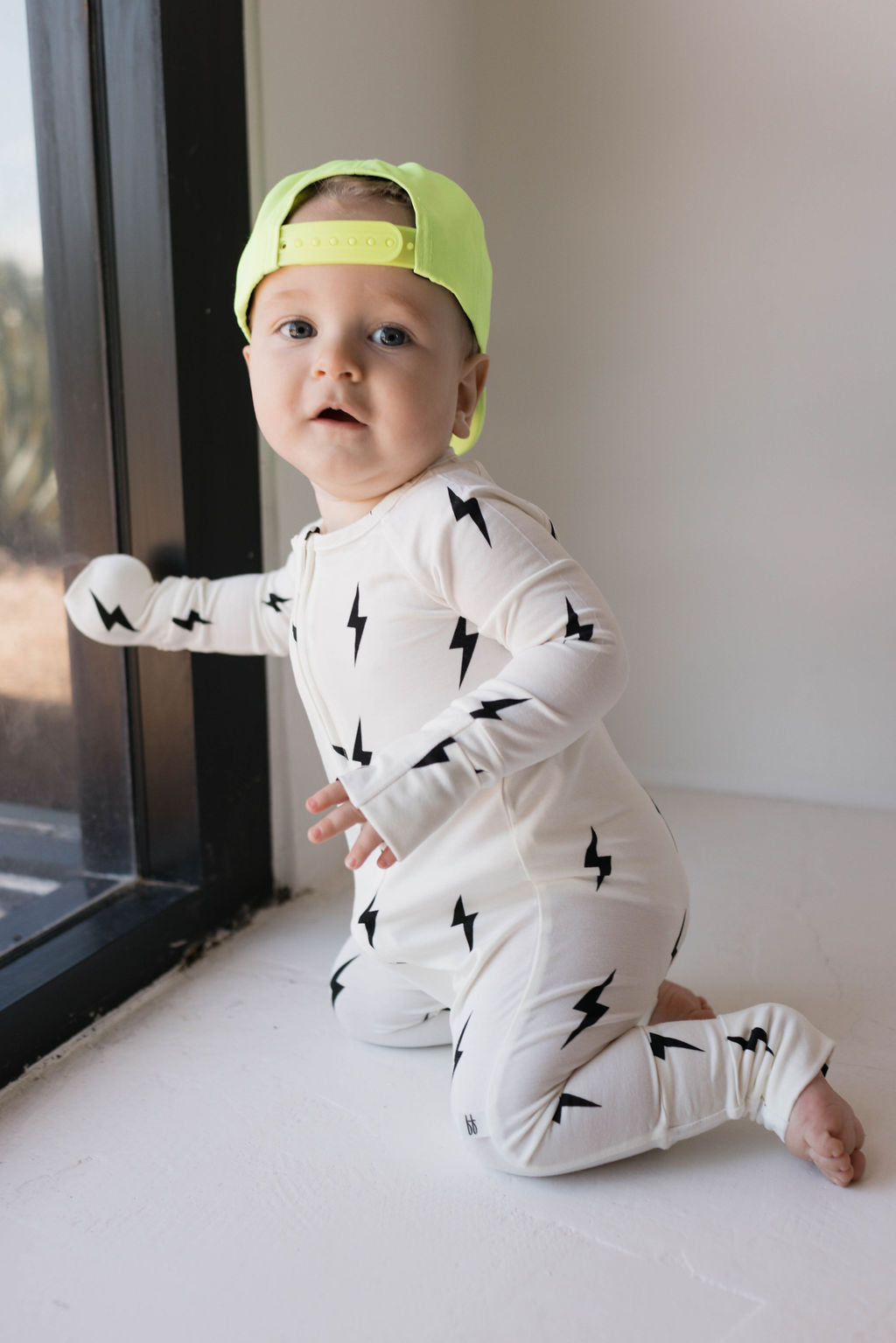 A baby wearing a "Children's Trucker Hat | Highlights" by forever french baby in neon green, with an adjustable fit, and a white onesie adorned with black lightning bolt patterns kneels on the floor, looking out a window.