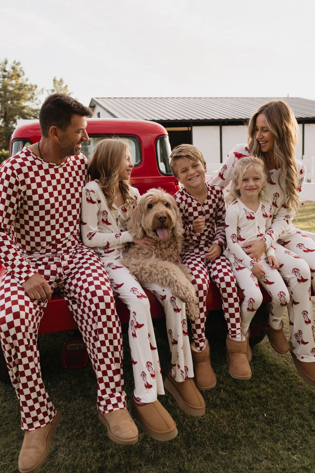 A family sits together on a red truck bed, all wearing matching Pre-Teen Flare Bamboo Pajamas in the Cowgirl Christmas pattern by lolo webb. The breathable fabric showcases charming red and white checkered designs. They are smiling with their large fluffy dog beside them, against a backdrop of a white building and trees.