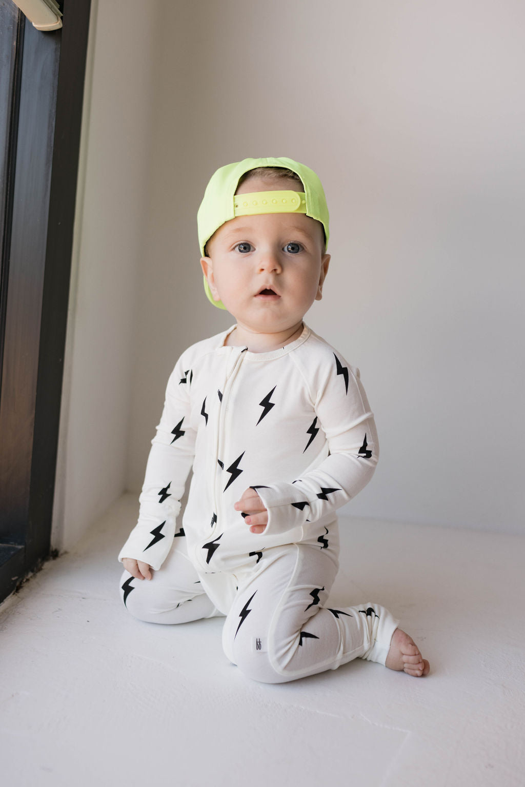 A baby sits on the floor next to a window, dressed in a white onesie adorned with black lightning bolt patterns. The baby sports the Children's Trucker Hat | Highlights by forever french baby, worn backward in neon yellow, and has a serious expression. Natural light streams in, illuminating the scene perfectly for future outdoor adventures.