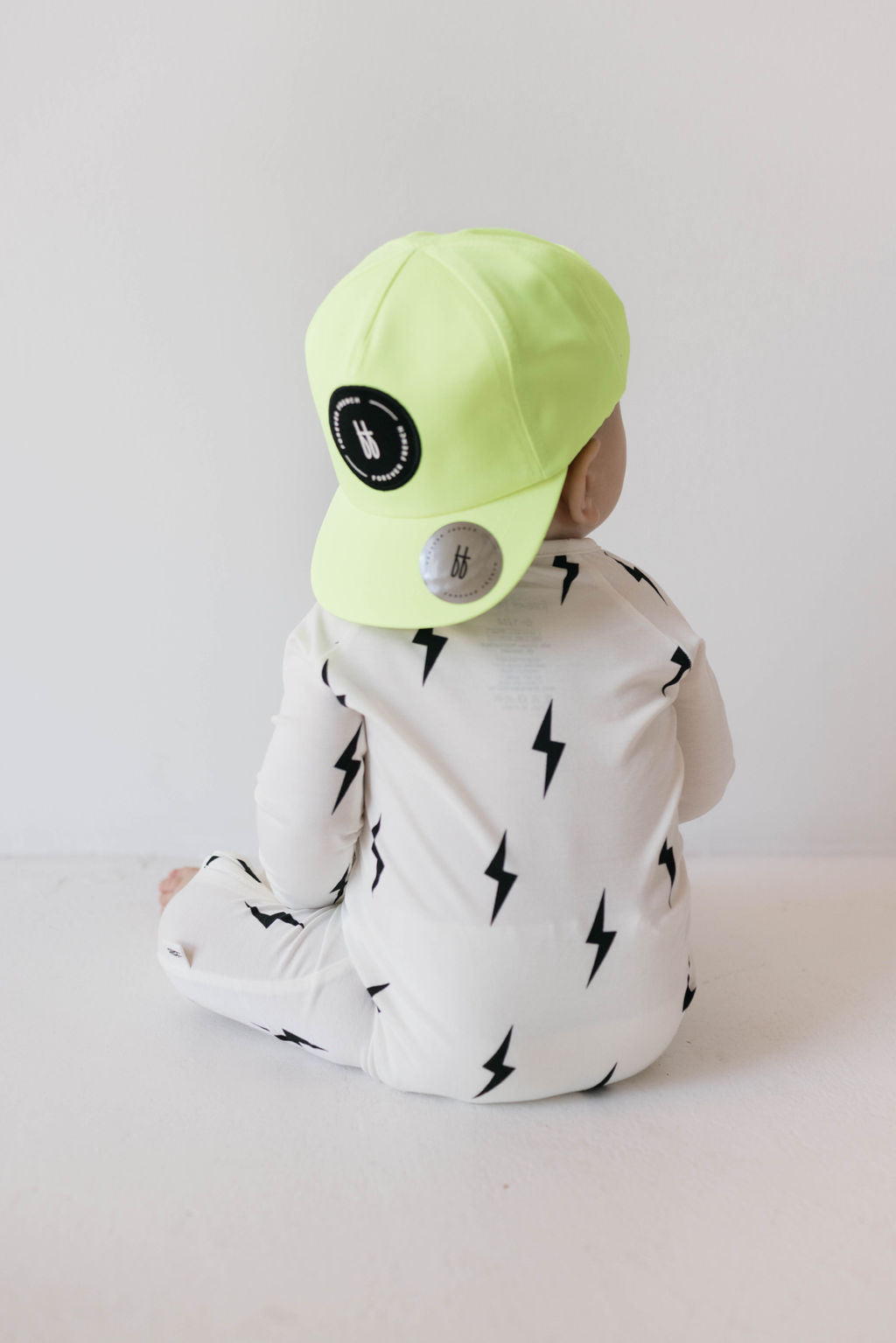 A baby is sitting on the floor wearing a cream-colored onesie adorned with black lightning bolts. The baby, facing away from the camera, is sporting a neon green Children's Trucker Hat from forever french baby. This hat features a black circular logo on the back and is designed for an adjustable fit, making it perfect for outdoor adventures. The background is plain white.