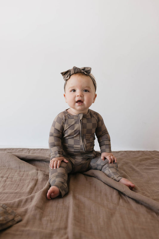 A baby dressed in a breathable, Faded Brown Checkerboard Bamboo Zip Pajamas from forever french baby, complete with a matching headband, is seated on a soft, brown blanket. The baby gazes slightly upward with a curious expression as light gently illuminates the simple, minimalist background.