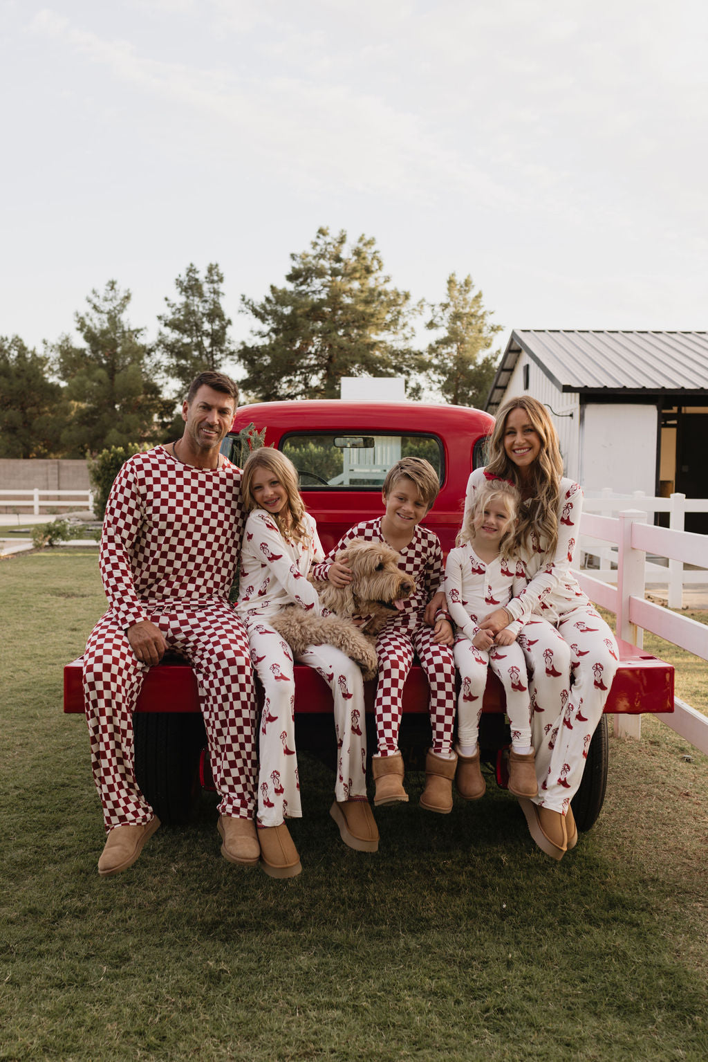 A family of five, beaming with smiles, relaxes on the back of a red truck wearing matching Pre-Teen Flare Bamboo Pajamas from lolo webb's Cowgirl Christmas collection. They're outdoors by a white fence and a barn, surrounded by trees. Their dog joins in the fun, reveling in the comfort of breathable fabric under the open sky.