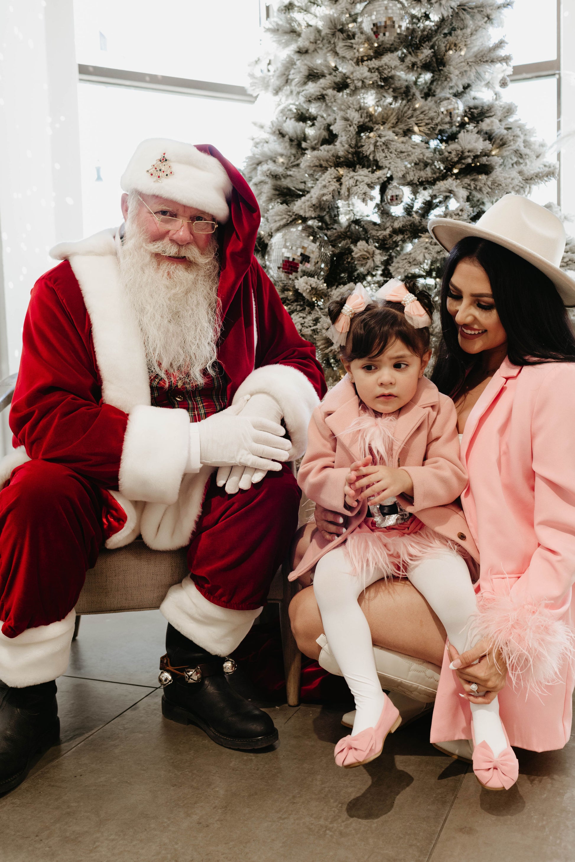 In a festive scene captured spectacularly by Amanda Riley Photos for the "Santa Photos | Forever French Baby x Amanda Riley Photos" product, a man dressed as Santa Claus sits in a chair beside a woman and a young girl, both wearing pink outfits. The girl sits on Santa's lap, with a decorated Christmas tree providing the perfect holiday backdrop.