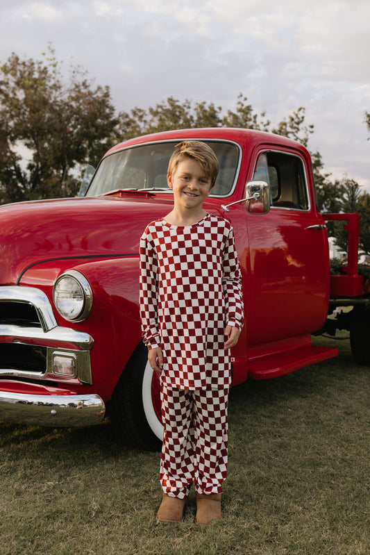 A young boy dressed in the Pre-Teen Straight Leg Bamboo Pajamas from forever french baby, known as the Quinn, stands smiling in front of a vintage red truck. The outdoor scene is set on a grassy area with trees and a partly cloudy sky, with his breathable pajamas capturing the joy of being in open air.