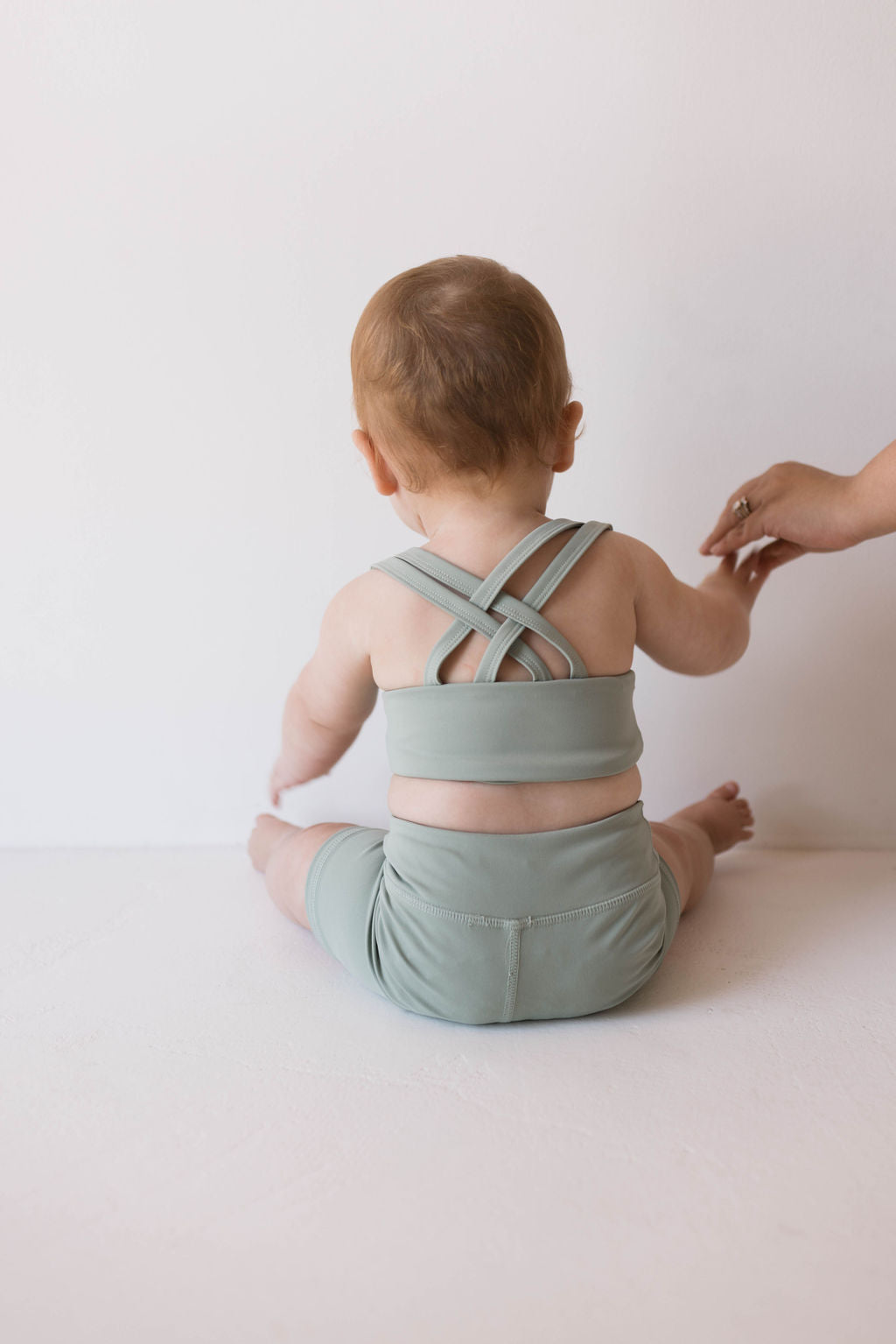 A baby sits on a light-colored surface, wearing the "Children's Workout Set | Green" by forever french baby, which includes a crisscross back top and shorts. An adult's hand gently touches the baby's arm, extended backwards against the plain, light-colored background.