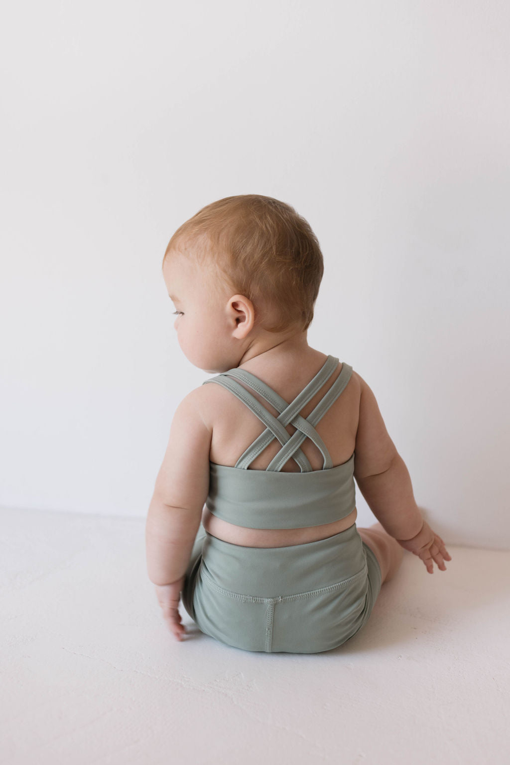 A baby wearing a green **Children's Workout Set | Green** from **forever french baby** sits on the floor facing away from the camera. The outfit features crisscross straps on the back. The baby's head is turned slightly to the left, with a light and simple background enhancing the minimalistic look.