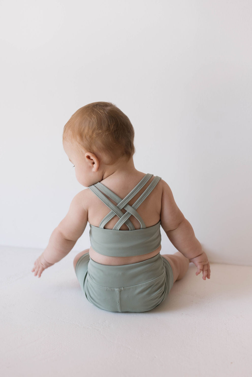 A baby with short light brown hair is sitting on a white surface facing away from the camera, wearing the Children's Workout Set in Green by forever french baby, which includes a crisscross back top and matching bottoms. The background is plain white.