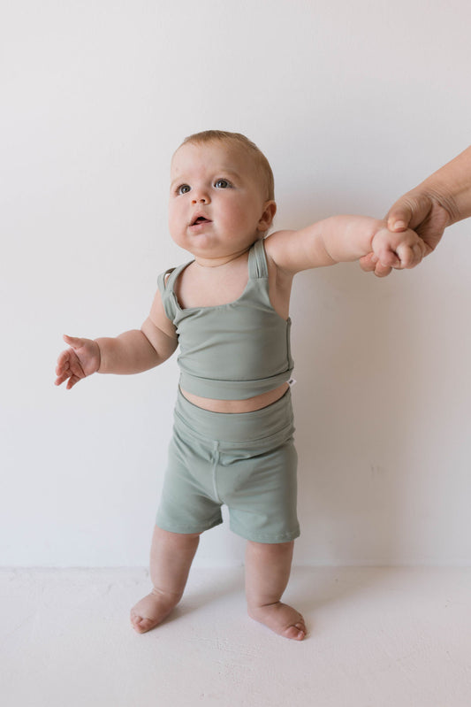 A baby wearing the Children's Workout Set in green from forever french baby stands on a white surface, holding onto an adult's hand for balance. The baby's eyes gaze upward and their mouth is slightly open, giving them an appearance of curiosity or surprise.