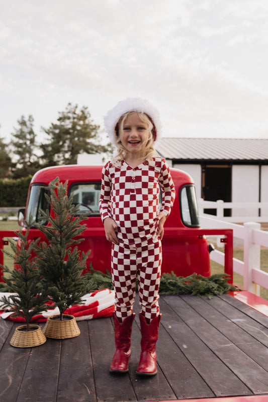 A child dressed in the Bamboo Two Piece Pajamas by lolo webb, featuring a red and white checkered pattern made of breathable fabric, and sporting a Santa hat, stands on a wooden platform. They are smiling in front of a classic red truck, surrounded by small evergreen trees and festive decor that create a holiday scene.