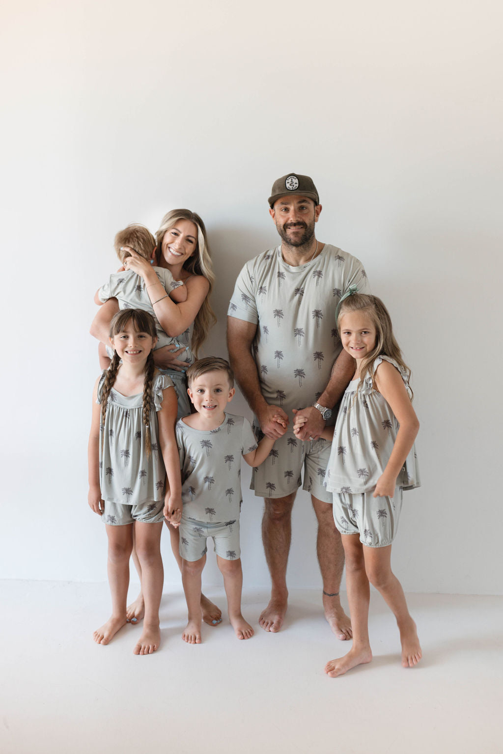 A family of six, dressed in matching Cami Women's Bamboo Set | Summer Dreamin' outfits from Forever French, stands together against a plain white background. The mother holds a baby while the father and three children stand beside them, all smiling. Their light gray attire, adorned with a palm tree print, looks comfortable and eco-friendly, made from hypo-allergenic bamboo fabric.