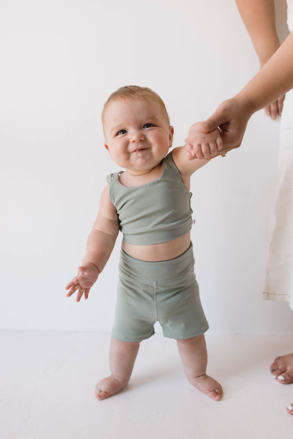 A smiling baby wearing the "Children's Workout Set | Green" by forever french baby stands while holding an adult’s hand. The baby appears happy and confident in a minimalistic setting with a plain background. The adult's torso is partially visible on the right side of the image.