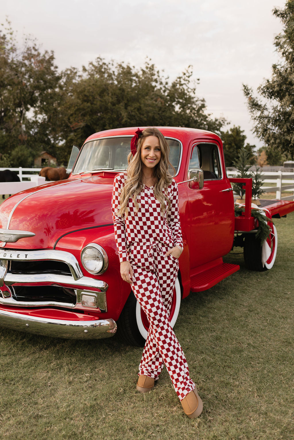 A person stands smiling in front of a vintage red truck, dressed in lolo webb's stylish Women's Bamboo Pajamas called the Quinn, featuring a charming red and white checkered design made from hypo-allergenic fabric. The truck is parked on grass with trees and a white fence in the background, creating a relaxed and cheerful atmosphere.