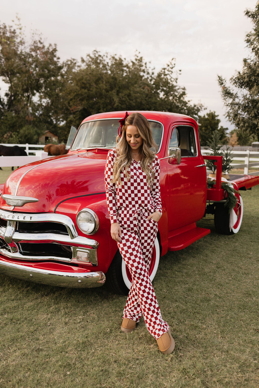 A woman clad in the Quinn pajamas by lolo webb, featuring a red and white checkered design, leans against a vintage red truck parked on a grassy lawn. Her cozy look stands out against the trees beneath a cloudy sky.