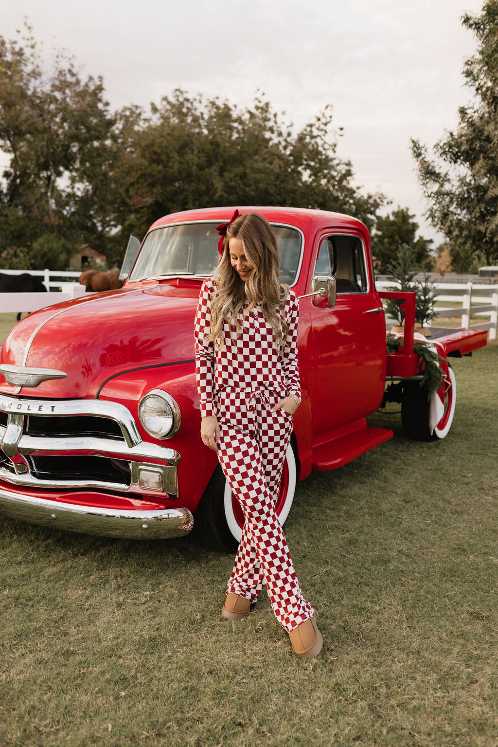 A woman in an outfit evoking the comfort and style of the Quinn bamboo pajamas by lolo webb stands next to a vintage red truck on a grassy area. The truck, featuring white wall tires and a wooden bed, is set against a backdrop of trees and a charming white fence.