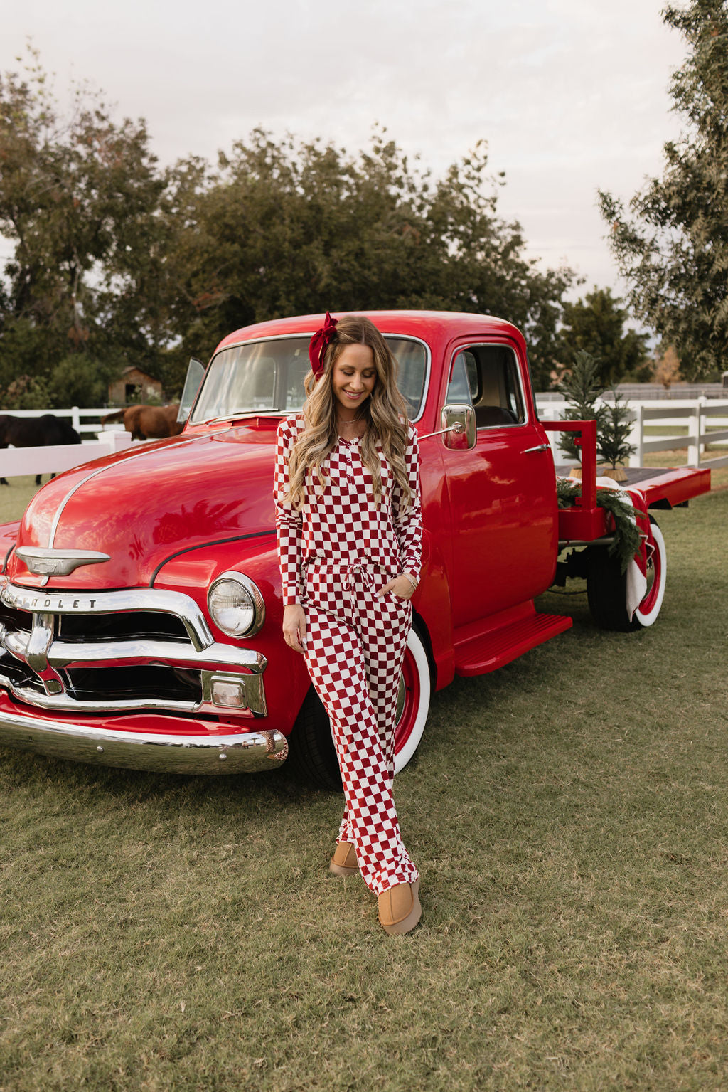 A woman dressed in the Quinn, the cozy red and white checkered bamboo pajamas from lolo webb, stands beside a vintage red truck on a grassy field. The serene backdrop of trees and a white fence enhances the charm of this hypo-allergenic clothing scene.