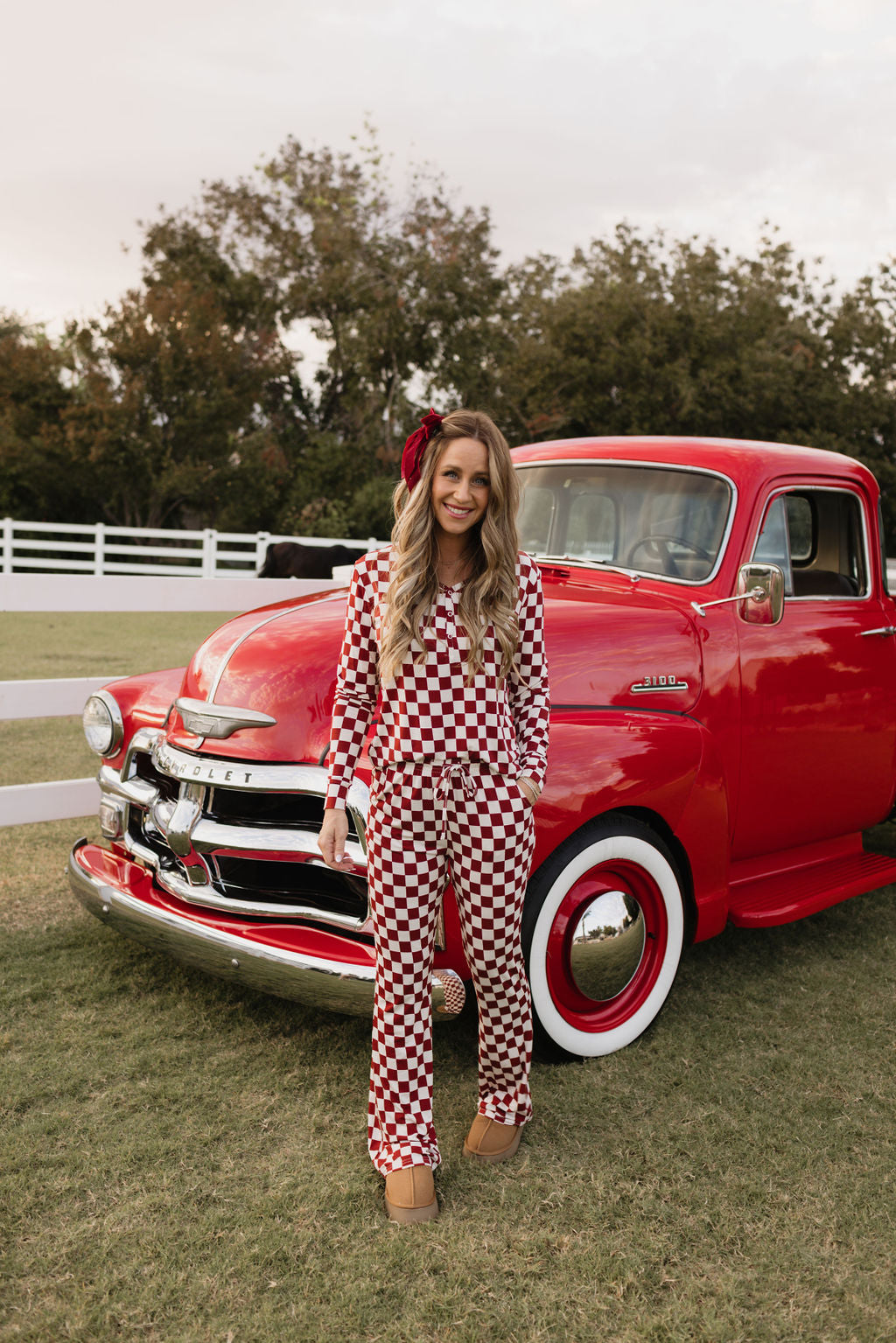 A person stands smiling in front of a vintage red truck, wearing the stylish red and white checkered Women's Bamboo Pajamas | the Quinn from lolo webb, made from hypo-allergenic materials. The scene is outdoors with a grassy field, white fence, and trees in the background.