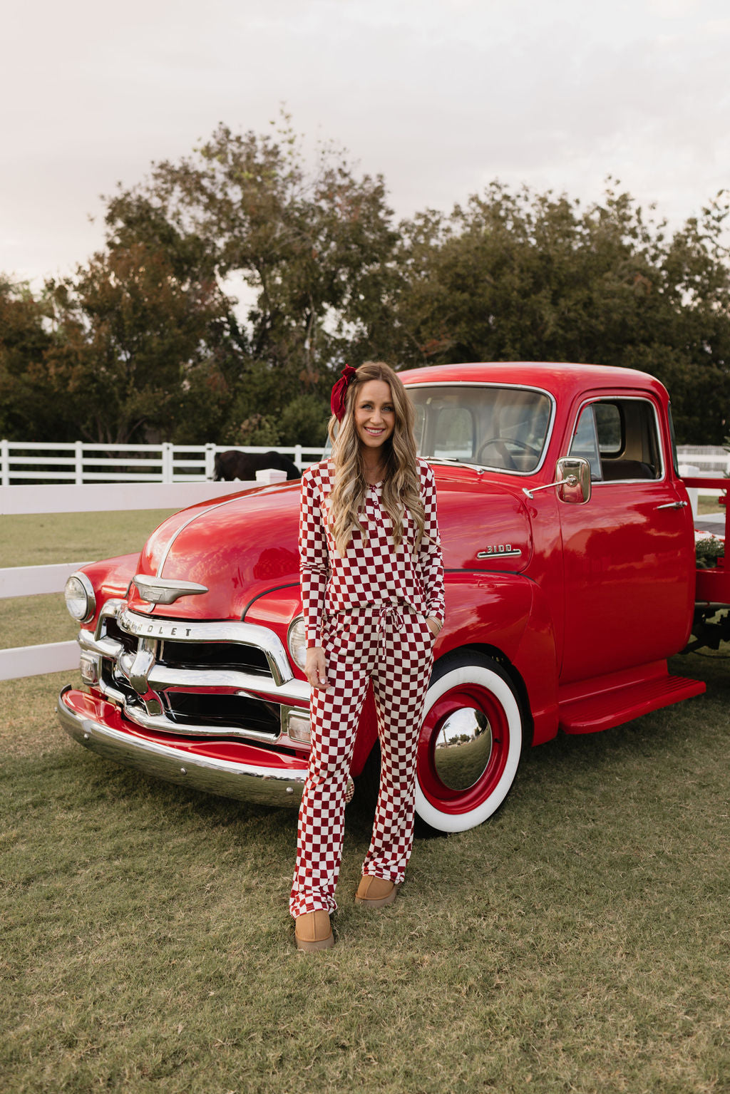 A woman wearing the lolo webb Women's Bamboo Pajamas, the Quinn model, featuring a red and white checkered design similar to breastfeeding pajamas, stands in front of a vintage red truck on a grassy field. Trees and a white fence create the backdrop.