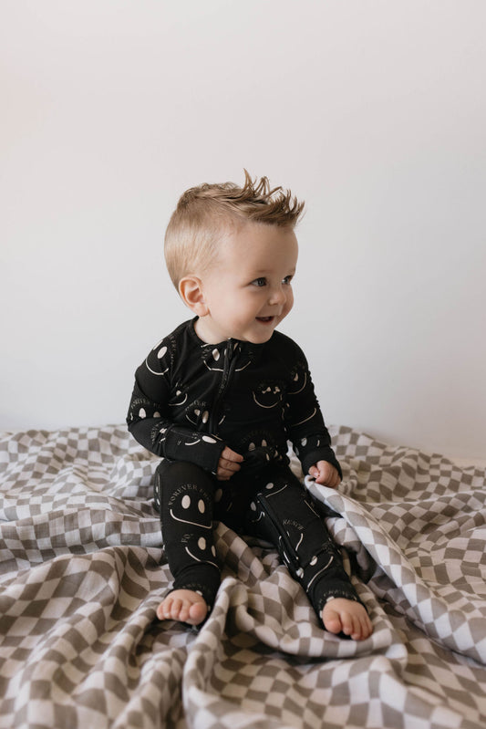 A happy baby with short blonde hair sits on a bed covered with a checkered blanket. The baby is wearing hypoallergenic, breathable Bamboo Zip Pajamas in Charcoal & White ff Smile from forever french baby. Smiling and looking off to the side, the baby is framed by a plain, light-colored wall in the background.