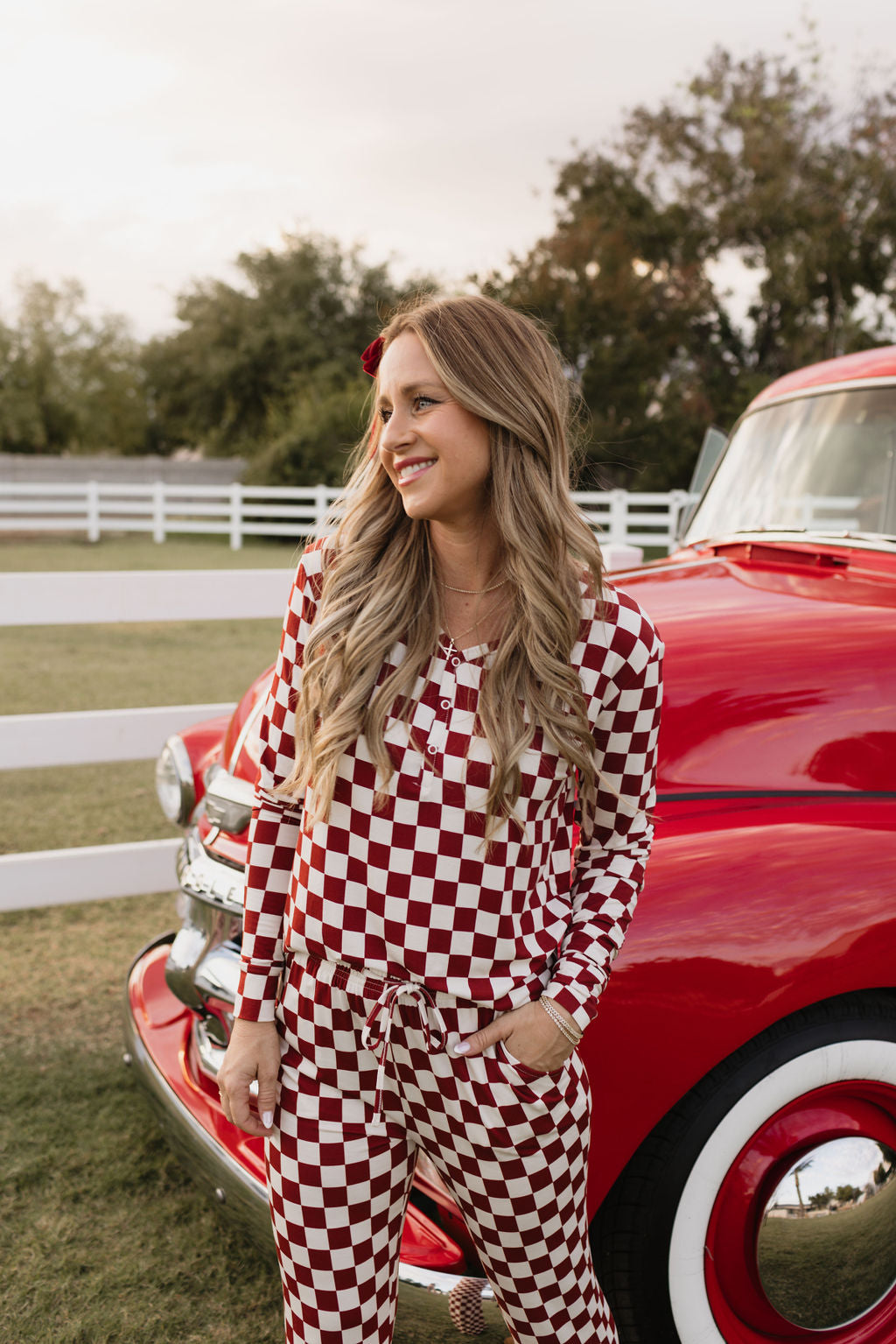 A person with long blonde hair stands smiling in front of a vintage red truck, dressed in the Quinn pajamas by lolo webb, which feature a cozy red and white checkered design. The scene is set against a backdrop of a white fence and trees beneath a partly cloudy sky.